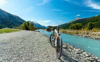 Weißes Fahrrad auf Schotterweg neben türkisfarbenem Fluss bei La Punt. Berge und Wälder rahmen das Tal unter blauem Himmel.