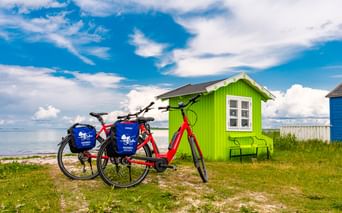 Zwei rote Fahrräder mit blauen Taschen vor einem hellgrünen Strandhaus auf Ærø. Im Hintergrund sind das Meer und der blaue Himmel mit Wolken sichtbar.
