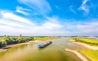 Luftaufnahme eines blauen Frachtschiffs auf dem Niederrhein bei Arnheim. Grüne Flussufer und städtische Gebäude unter blauem Himmel.