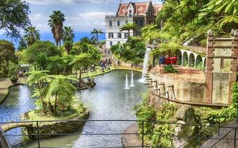 Terrassierter tropischer Garten in Funchal, Madeira mit Teichen, Springbrunnen, üppiger Vegetation und weißem Palast mit rotem Dach.