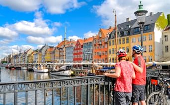 Zwei Radfahrer mit Helmen betrachten bunte historische Gebäude entlang des Nyhavn-Hafens in Kopenhagen. Traditionelle Boote liegen im Kanal.
