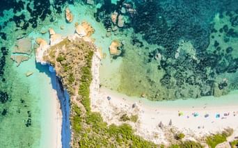 Luftaufnahme der Badebucht Padulella auf Elba mit weißem Sand, türkisfarbenem Wasser, felsiger Küste, grüner Vegetation und bunten Sonnenschirmen.