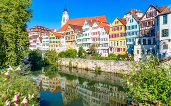 Row of colorful half-timbered houses along the Neckar River in Tübingen, with a church tower and reflections in the water, framed by green foliage.