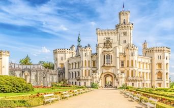 Weißes Schloss Hluboka mit zinnenbesetzten Türmen und gotischer Architektur. Ein Kiesweg führt durch formale Gärten mit Hecken.