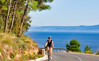 Radfahrerin mit Helm auf Küstenstraße in Kroatien. Blaues Meer und ferne Berge sichtbar, Kiefern säumen den Hang.