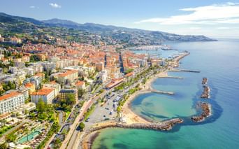 Panoramablick auf Sanremo, Italien, mit Küstenstadt, Stränden, Hafen, palmengesäumter Promenade und Bergen im Hintergrund.