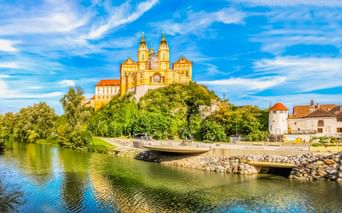 Yellow baroque Melk Abbey with twin towers sits atop a green hill above the Danube River. A curved promenade lines the riverbank under blue sky.