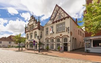 Fachwerkgebäude mit weißer Fassade der Apotheke am Marktplatz in Tönning. Traditionelle deutsche Architektur mit Kopfsteinpflaster unter blauem Himmel.