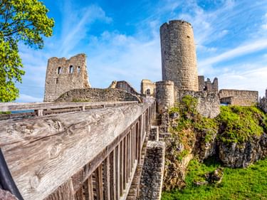 Wolfstein Castle ruins in Neumarkt with a tall round tower and stone walls. A wooden walkway leads to the ruins under a blue sky with clouds.