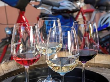 Fünf Weingläser mit Rosé-, Weiß- und Rotwein auf einem schwarzen Tablett. Rote Fahrräder sind im Hintergrund in Maikammer sichtbar.
