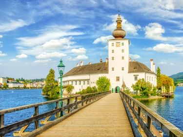 Holzsteg führt zu Schloss Orth, einem weißen Schloss mit Turm auf einer Insel im Traunsee bei Gmunden, Österreich, unter blauem Himmel.