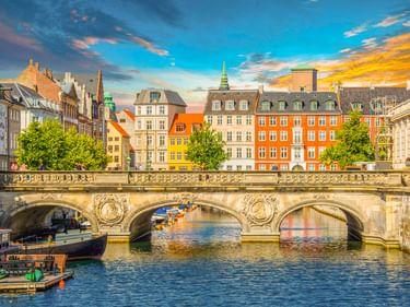 Steinbrücke über Kanal mit bunten historischen Gebäuden in Kopenhagens Altstadt. Grüner Kirchturm im Hintergrund unter dramatischem Abendhimmel.