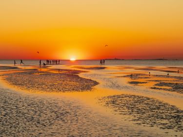 Goldener Sonnenuntergang über dem Watt bei Cuxhaven und der Insel Neuwerk. Silhouetten von Menschen auf nassem Sand mit Wasserpfützen.