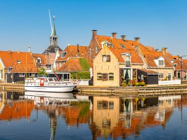 Weißes Segelboot am Kanal in Makkum mit traditionellen niederländischen Häusern mit orangen Ziegeldächern und Kirchturm, gespiegelt im Wasser.