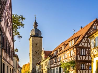 Historic Hexenturm tower with domed roof rises above traditional half-timbered houses with orange tile roofs in Zeil am Main under blue sky.