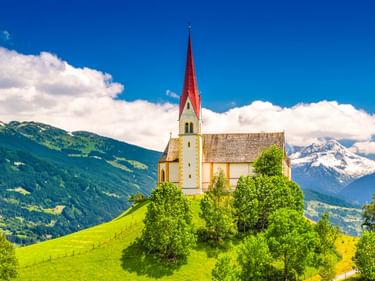 Weiße Kirche mit rotem Turm auf grünem Hügel in den österreichischen Alpen. Berge und blauer Himmel mit Wolken im Hintergrund.