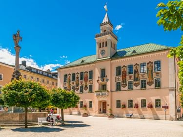 Historisches Rathaus in Bad Reichenhall mit bemalter Fassade, Uhrturm und grünem Kupferdach. Eine Statuensäule steht auf dem Platz mit Bäumen.
