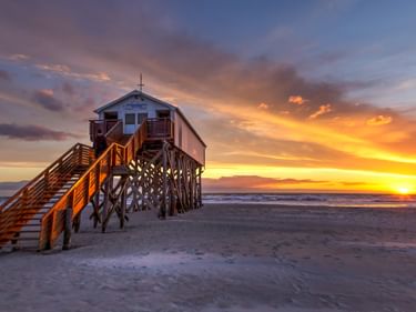 Hölzerner Pfahlbau mit Treppe am Strand von Sankt Peter-Ording bei Sonnenuntergang. Orange und violetter Himmel über dem Nordseestrand.