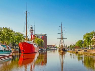 Rotes historisches Schiff im Emder Hafenkanal mit hohen Segelschiffen im Hintergrund. Bäume säumen das Ufer unter blauem Himmel.