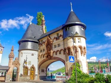 Historisches Brückentor mit zwei Rundtürmen und Kegeldächern in Traben-Trarbach unter blauem Himmel. Weinberge auf Hügeln im Hintergrund.