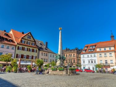 Historischer Marktplatz in Kulmbach mit bunten Gebäuden, Kopfsteinpflaster, zentralem Denkmal mit goldenem Stern und Straßencafé unter blauem Himmel.