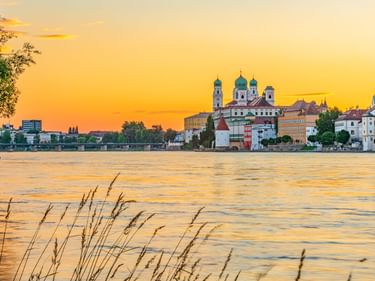Passauer Stadtansicht bei Sonnenuntergang mit Domtürmen und historischen Gebäuden an der Donau unter orangefarbenem Himmel.