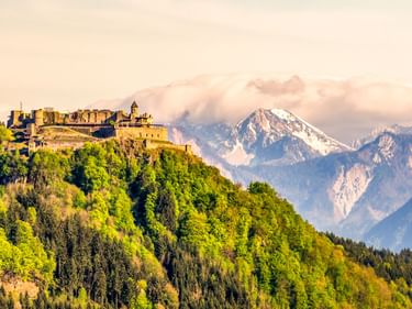 Burg Landskron castle perched on a forested hilltop near Villach, with snow-capped Alpine peaks visible in the background under a soft sky.