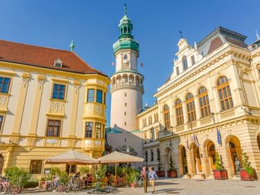 Historischer Feuerturm mit grüner Kuppel am Hauptplatz von Sopron, umgeben von gelben Barockgebäuden. Radfahrer und Fußgänger genießen Straßencafés.