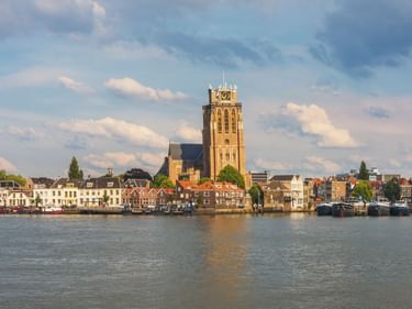 Blick auf die Dordrechter Uferpromenade mit hohem Kirchturm. Historische Gebäude säumen den Hafen mit Booten am Wasser.