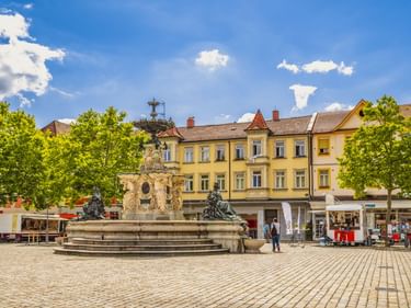Schlossplatz in Erlangen mit prächtigem Barockbrunnen auf Kopfsteinpflaster, gelben historischen Gebäuden, grünen Bäumen und blauem Himmel.