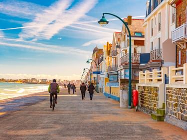 Radfahrer und Fußgänger auf der Uferpromenade in Saint-Malo. Bunte Häuser säumen den Weg, links ist das Meer unter blauem Himmel sichtbar.