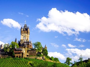 Reichsburg Cochem mit Turm und Türmchen auf einem Hügel über grünen Weinbergen unter blauem Himmel mit weißen Wolken an der Mosel.