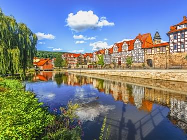 Half-timbered houses with red roofs reflected in calm river water in Hannoversch Münden. Stone wall and willow tree frame the waterfront scene.