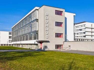 Das ikonische Bauhaus-Gebäude in Dessau mit weißer Fassade, Glasvorhangfassade und vertikalem BAUHAUS-Schriftzug. Rote Akzente an Balkonen.