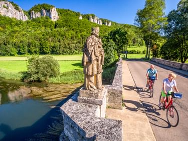 Two cyclists crossing a stone bridge with a statue in Hausen im Tal. White limestone cliffs and green forest visible in background under blue sky.