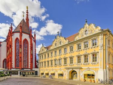 Red and white Gothic Marienkapelle church beside ornate yellow Rococo Haus zum Falken building on market square in Würzburg under blue sky.
