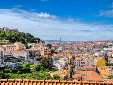 Panoramablick auf Lissabon mit orangefarbenen Ziegeldächern, bunten Gebäuden und einer Hängebrücke in der Ferne unter blauem Himmel.