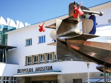 Weißes modernistisches Gebäude mit Zeppelin Museum Schriftzug und großer Metall-Propeller-Skulptur mit roten Elementen am Eingang in Friedrichshafen.