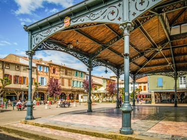 Verzierte Eisen- und Holzmarkthalle in Mirepoix mit dekorativen Säulen und grünem Metallwerk. Bunte Fachwerkhäuser säumen den Platz.