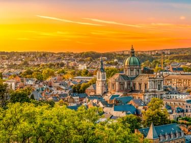 Panoramablick auf Namur, Belgien bei Sonnenuntergang mit der grünen Kuppel und dem Glockenturm der Kathedrale inmitten von Wohngebäuden und Bäumen.