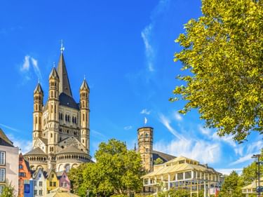 St. Martin Church in Cologne with four towers and dark spire against blue sky, surrounded by colorful historic buildings and green trees.