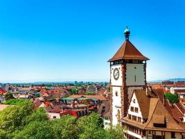 Historisches Schwabentor mit Uhr und rotem Dach in Freiburg, umgeben von roten Ziegeldächern und grünen Bäumen unter blauem Himmel.