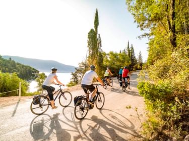 Gruppe von Radfahrern auf einer sonnigen Küstenstraße auf den Ionischen Inseln mit Meerblick, Zypressen und üppiger grüner Vegetation.