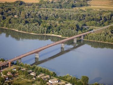 Luftaufnahme einer langen Brücke über die Donau bei Batina, mit dichten Wäldern an beiden Ufern und landwirtschaftlichen Feldern im Hintergrund.