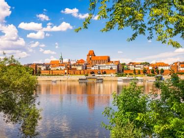 Historische Skyline von Toruń, Polen, mit roter Backstein-Kathedrale und Altstadtgebäuden, die sich in der Weichsel spiegeln, eingerahmt von grünen Bäumen.