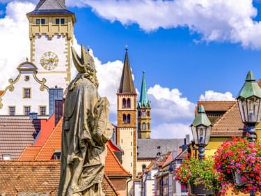 Würzburger Altstadt mit Statue, Uhrturm, Kirchtürmen und bunten Blumenkörben. Traditionelle Gebäude mit orangen Dächern unter blauem Himmel.