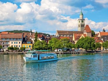 Blaue Personenfähre auf dem Bodensee vor der Uferpromenade von Überlingen. Historischer Kirchturm und bunte Gebäude unter blauem Himmel mit Wolken.