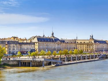 Panoramablick auf die Uferpromenade von Bordeaux mit klassischer Architektur entlang der Garonne. Bäume säumen die Promenade unter blauem Himmel.