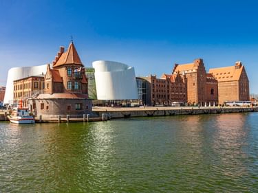 Blick auf den Stralsunder Hafen mit dem modernen weißen Ozeaneum-Gebäude neben historischen Backsteinbauten unter blauem Himmel.
