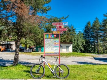 Weißes Fahrrad vor einer roten Informationstafel mit Radwegschildern und Karte in Scuol, umgeben von Bäumen und Gebäuden.